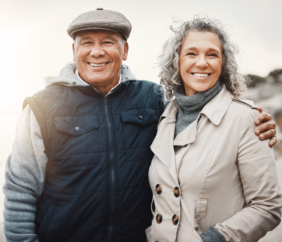 Smiling senior couple outdoors with the man wearing a gray cap and navy vest, and the woman in a beige coat and gray turtleneck.